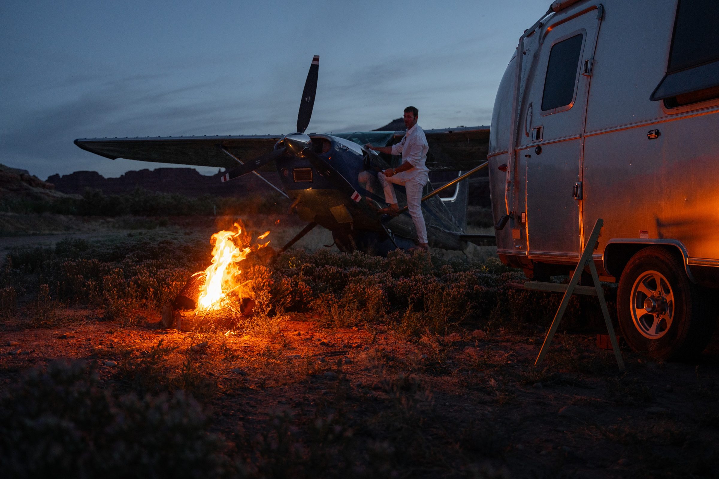 A campfire beside an Airstream and a parked aircraft under a night sky