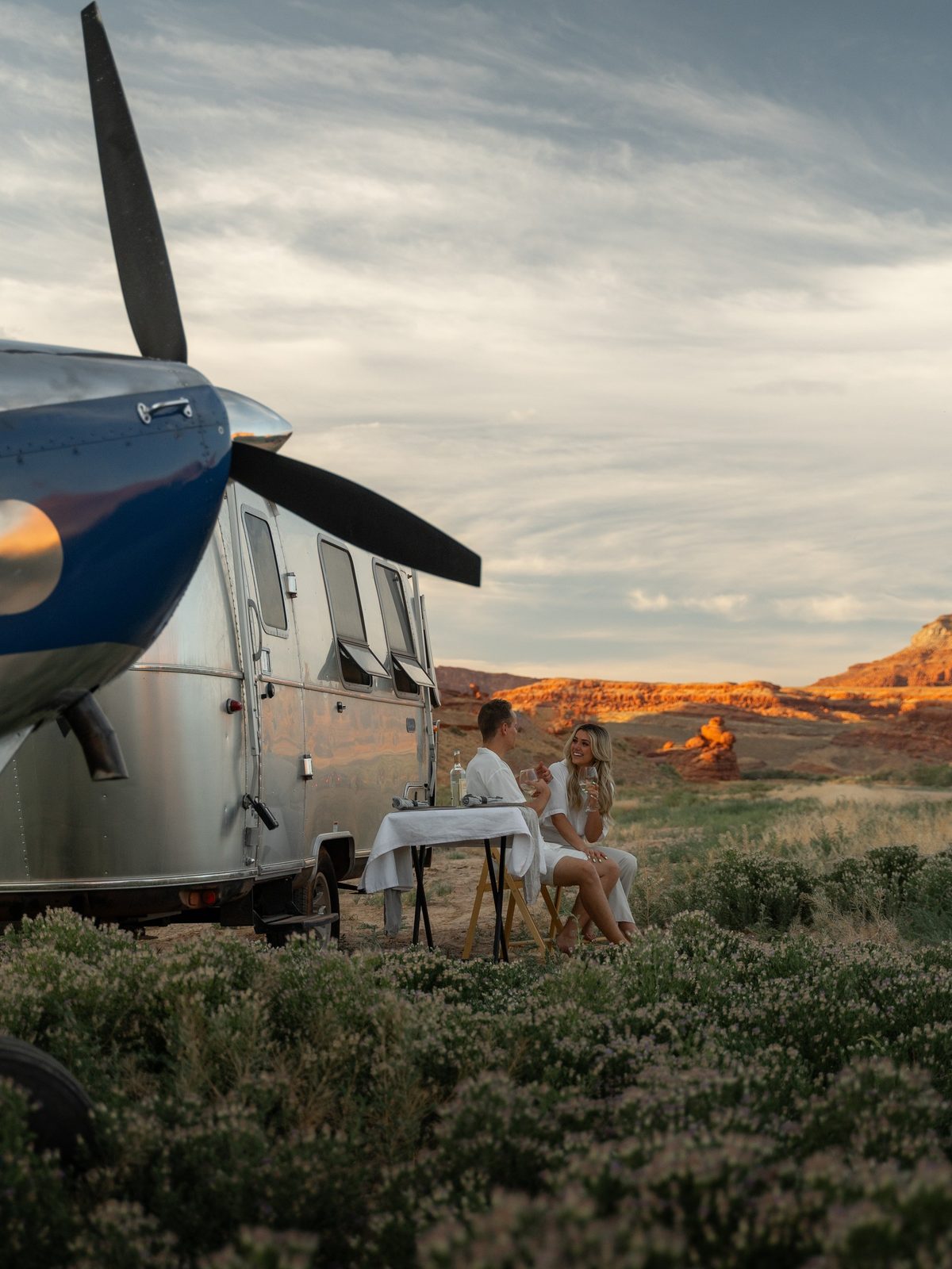 A dinner table set between an Airstream and a parked aircraft in warm evening light