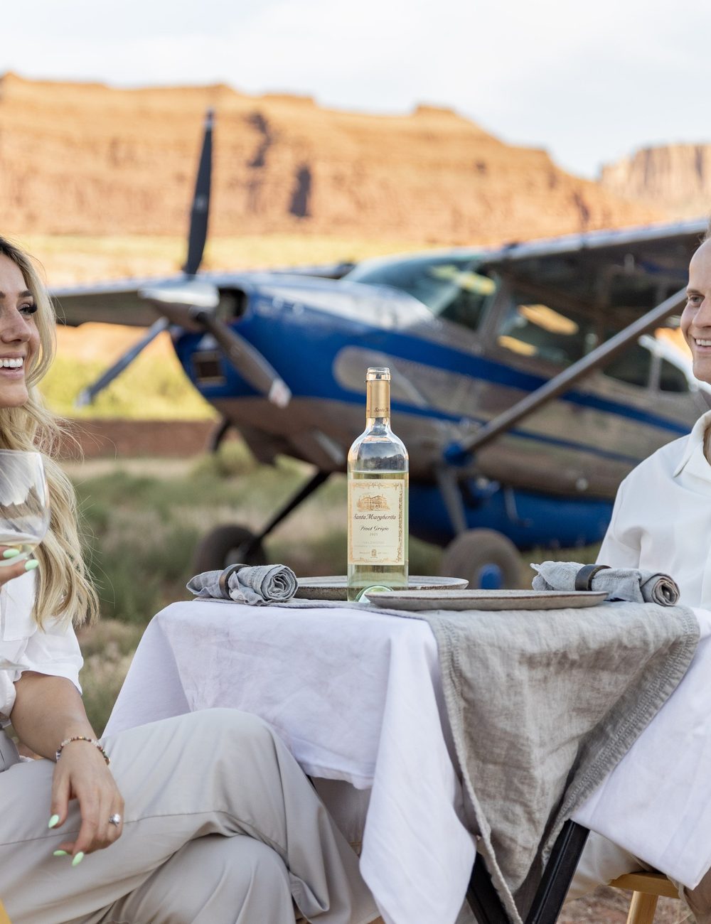 Two guests at a dinner table set beside a parked aircraft below red cliffs